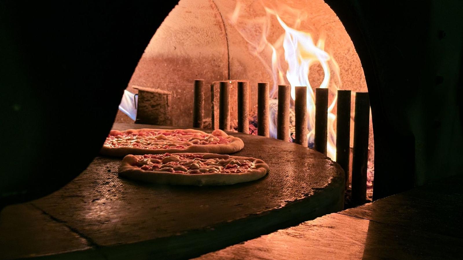Zwei Pizzen im Holzofen, dahinter lodern die Flammen