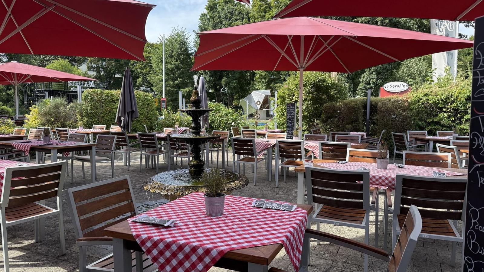 Terrasse mit plätscherndem Brunnen, roten Sonnenschirmen und rot-weiß karierten Tischdecken